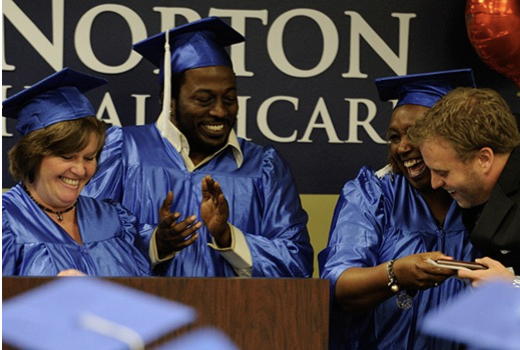 Graduates in blue gowns celebrate on stage, clapping and smiling behind a podium.