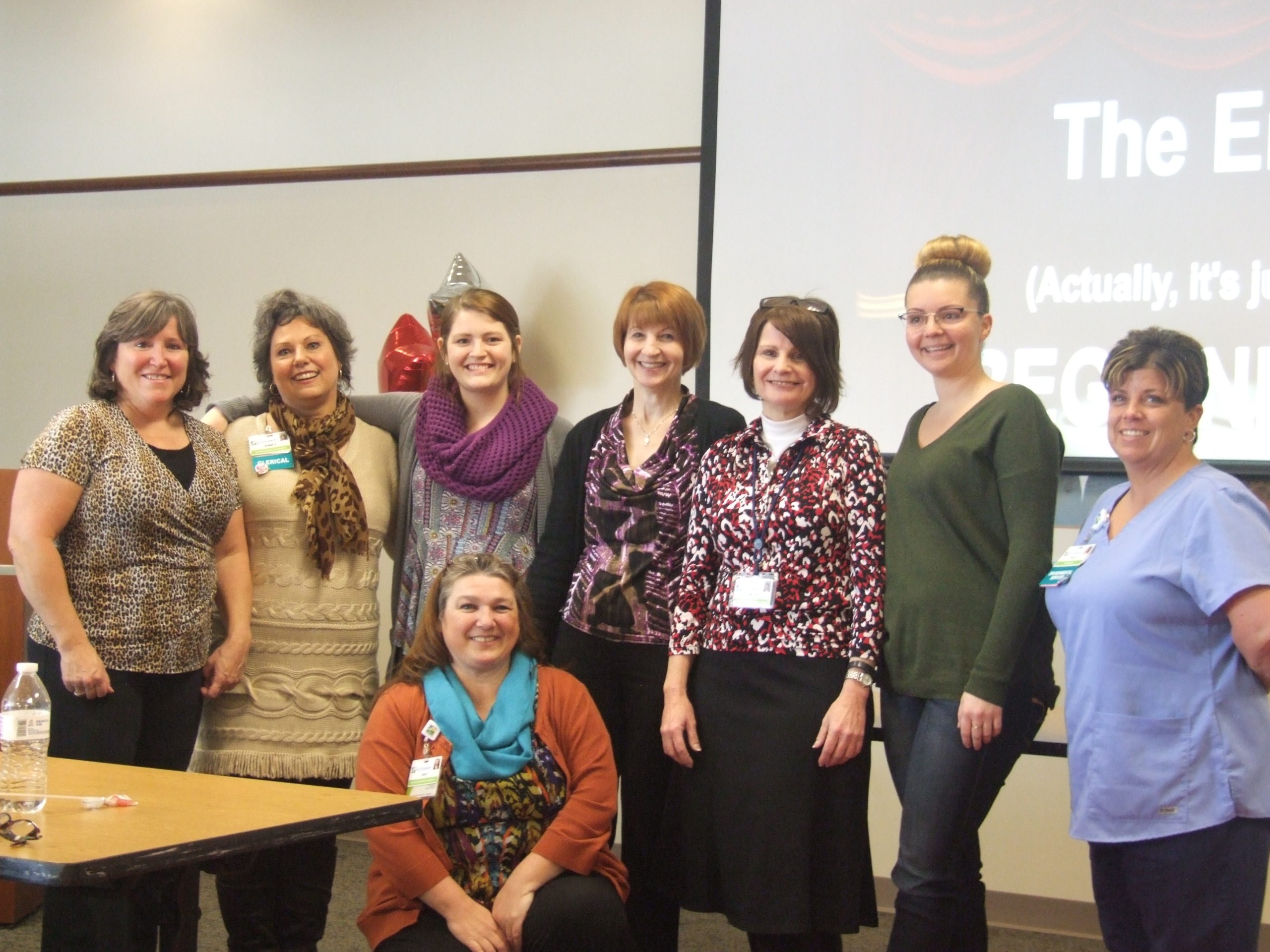 Group of nine women posing together in a meeting room, many wearing badges and colorful scarves, smiling at the camera.
