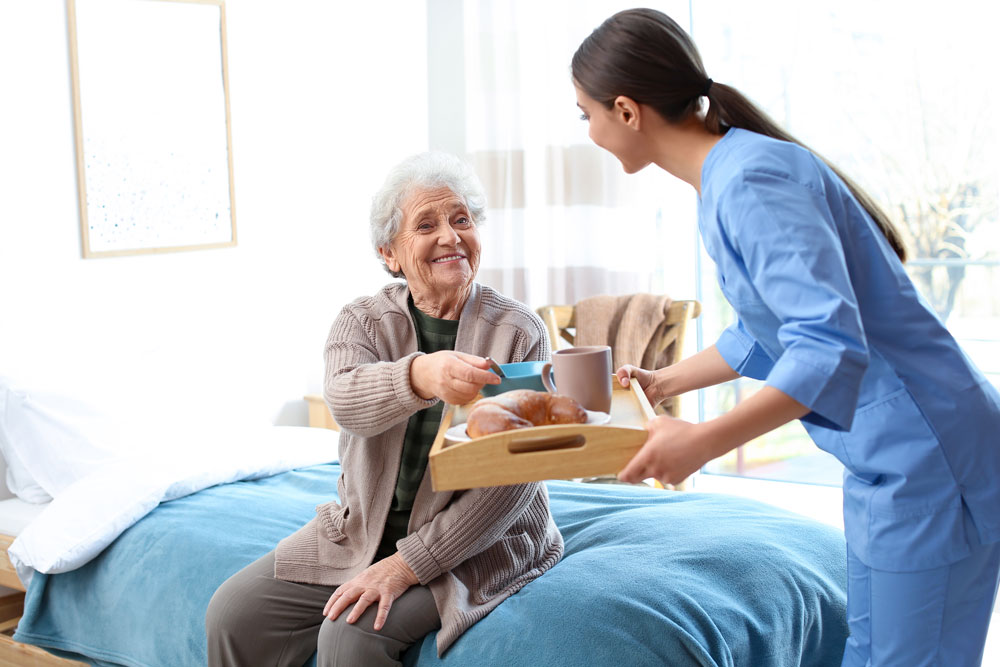 Elderly woman in a beige cardigan sits up in bed, smiling as a caregiver in blue scrubs brings a breakfast tray with coffee and pastries.