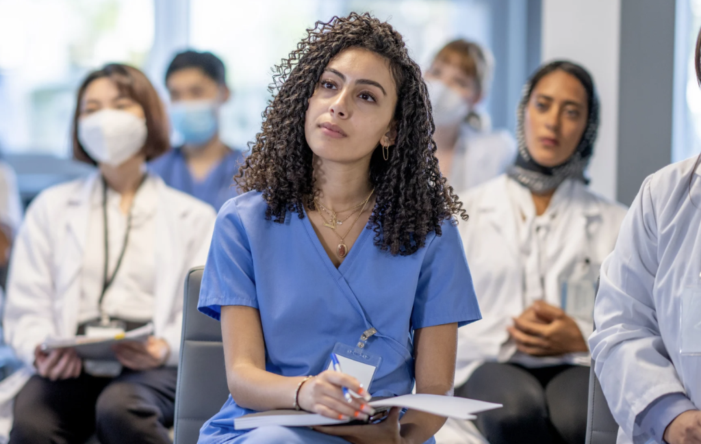 Front-and-center healthcare professional in blue scrubs with curly hair taking notes during a meeting, with other clinicians in the background